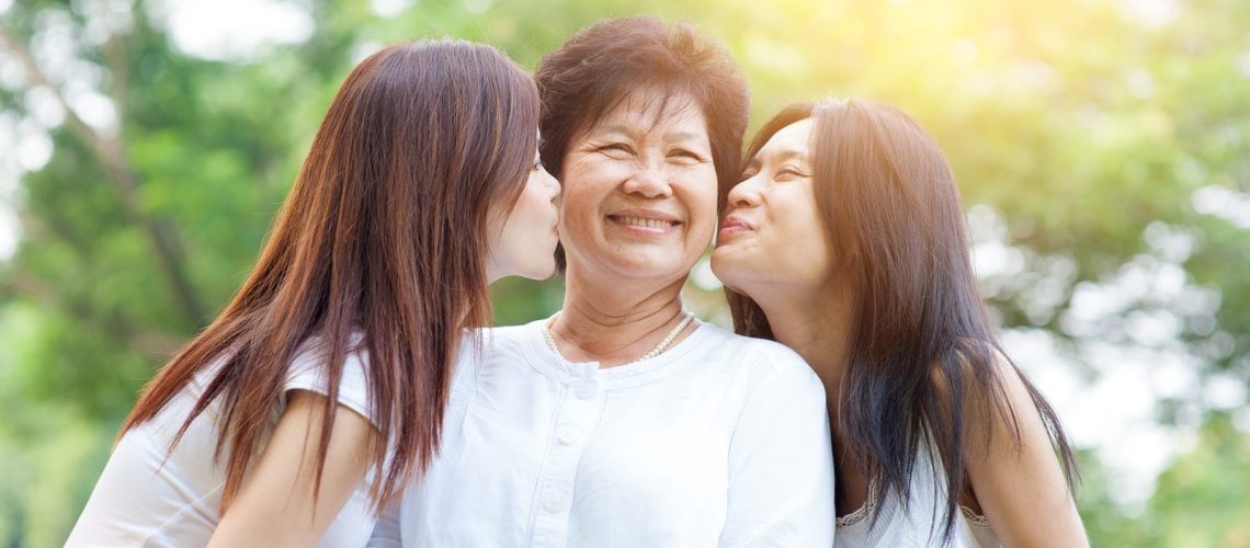 Portrait of Asian daughters kissing elderly mother, senior adult woman and grown child. Outdoors family at nature park with beautiful sun flare.