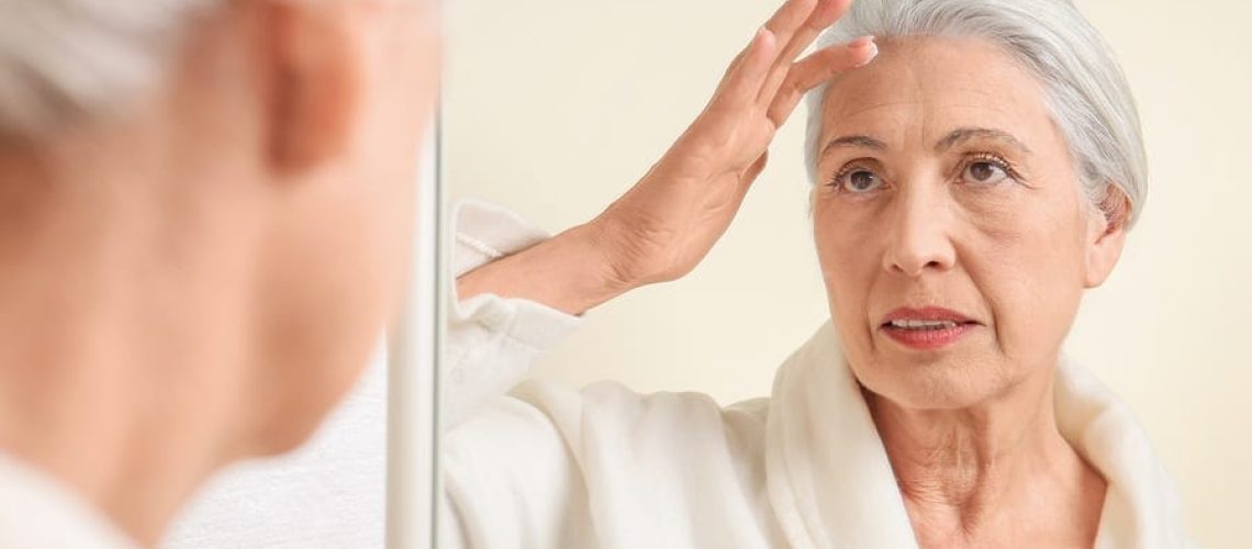 Beautiful elderly woman applying face cream in front of mirror at home